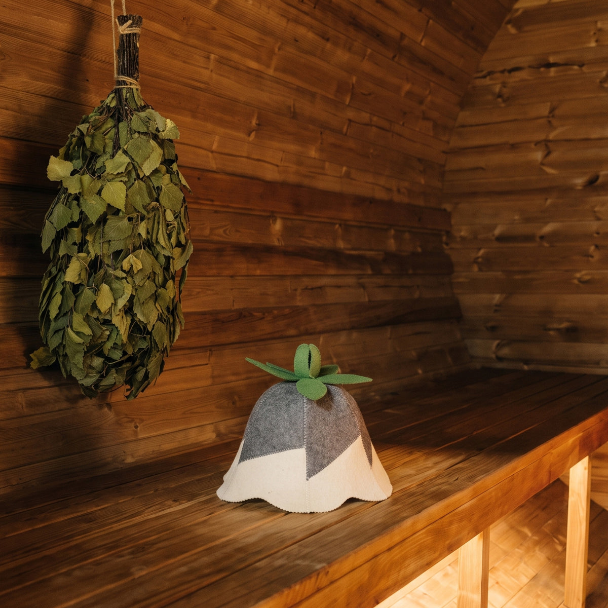 A small felted wool bell-shaped item with a gray and white pattern and green leaf detail sits on a wooden sauna bench, with a hanging bundle of green leaves against a wood-paneled wall.
