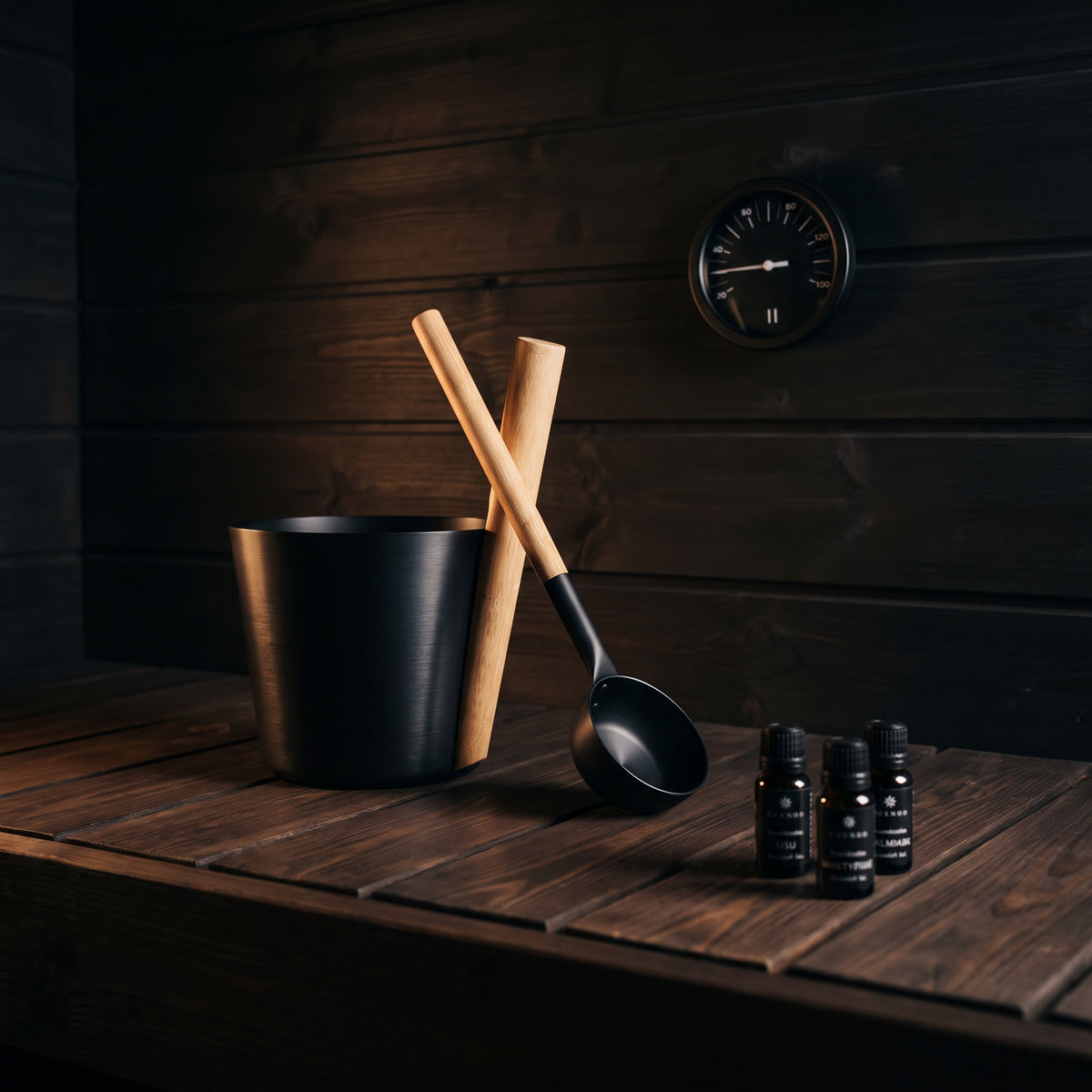 A black metal sauna bucket with two wooden-handled ladles resting inside, alongside three small black bottles, set on a dark wooden table against a dark wood-paneled wall.