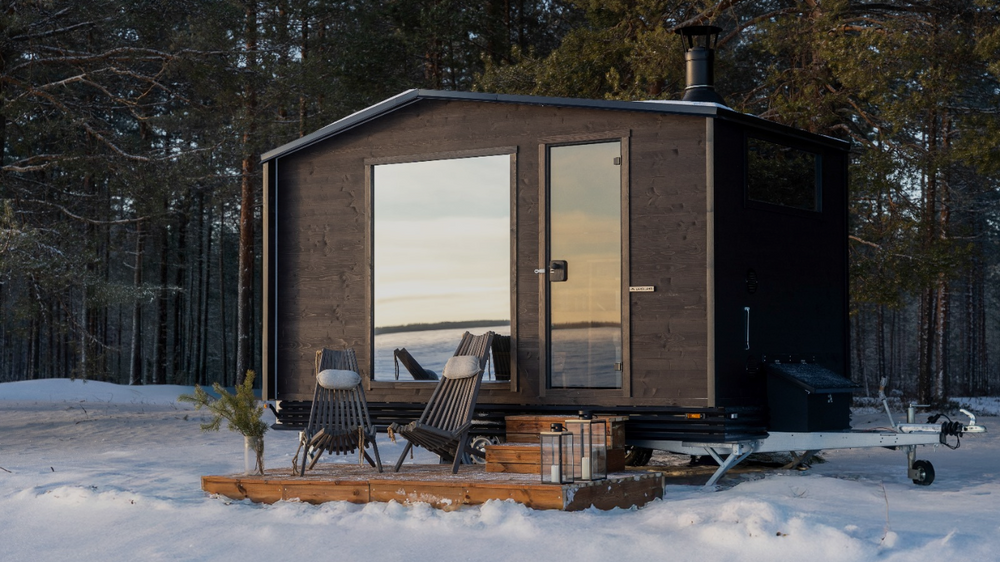 Small wooden cabin in a snowy forest with chairs and a table outside.