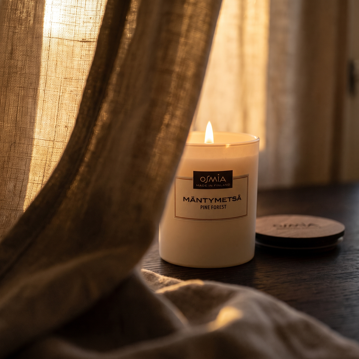 Pine Forest (Mäntymetsä) Scented Candle by Osmia - lit behind linen curtain