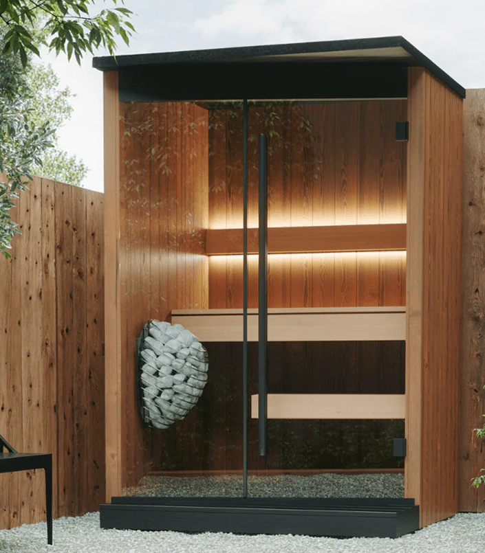 Wooden outdoor sauna with a black roof and interior lighting, surrounded by greenery.